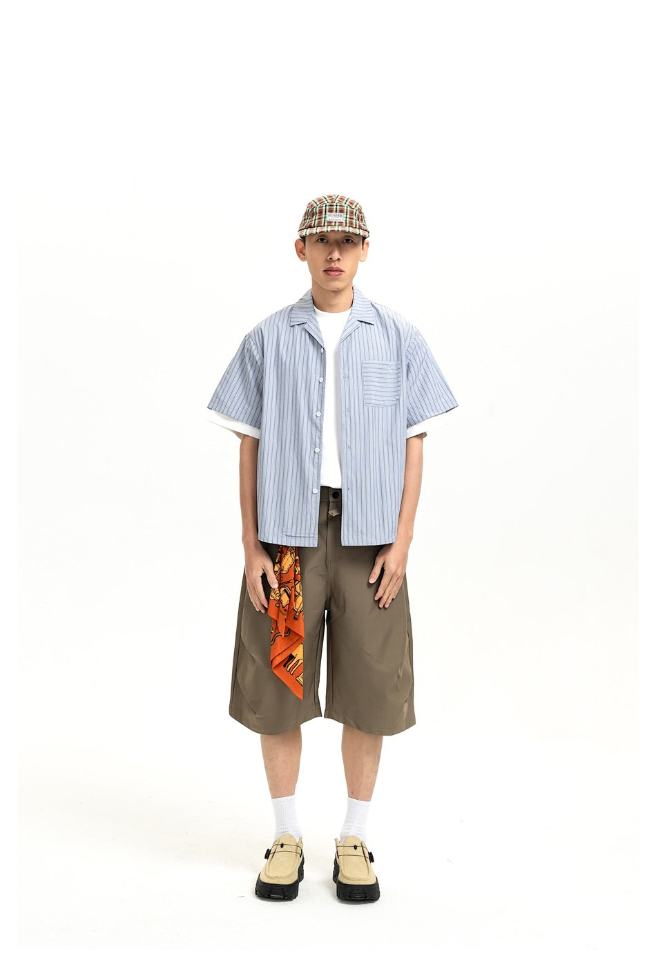Man wearing a blue striped short-sleeve camp collar shirt open over a white tee with olive shorts and a plaid cap