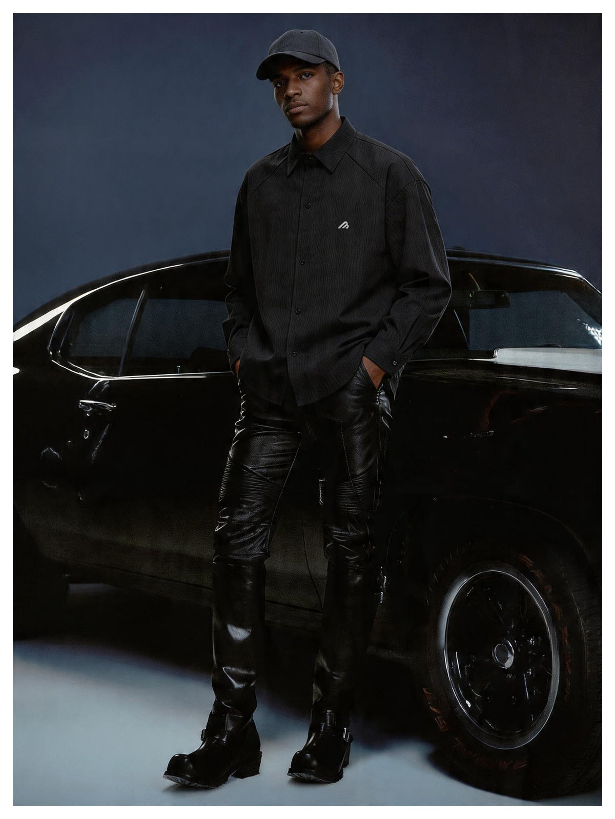 Man wearing a black textured-stripe button-up shirt with a cap, black leather pants and boots, standing by a black car