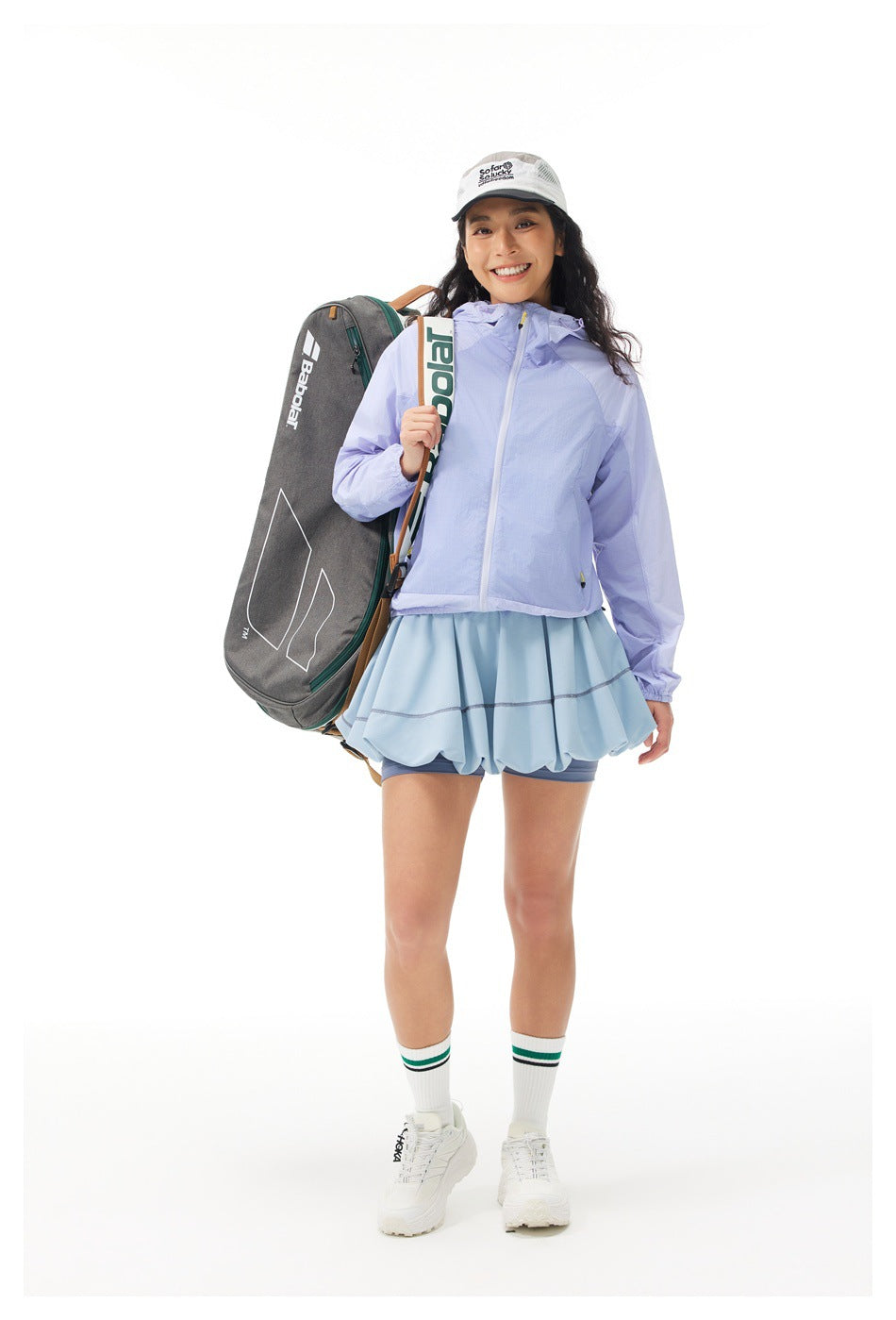 Woman in a lavender hooded jacket and pale blue ruffled athletic skirt, carrying a tennis racket bag.