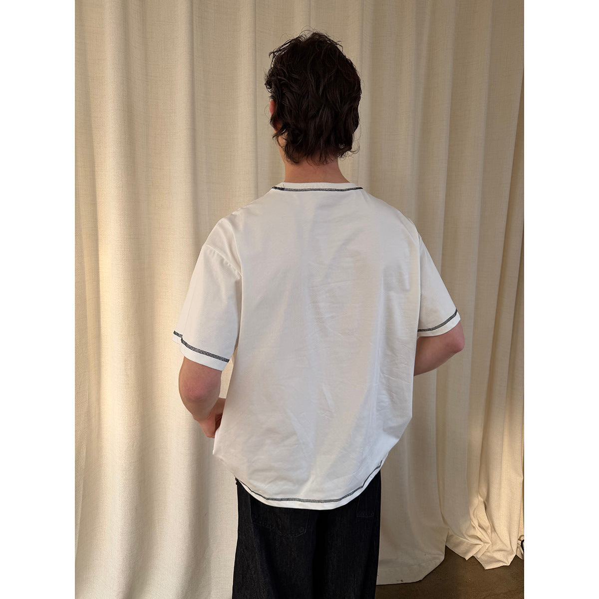 Back view of a model wearing a white T-shirt with black contrast stitching at the collar, sleeves, and hem.