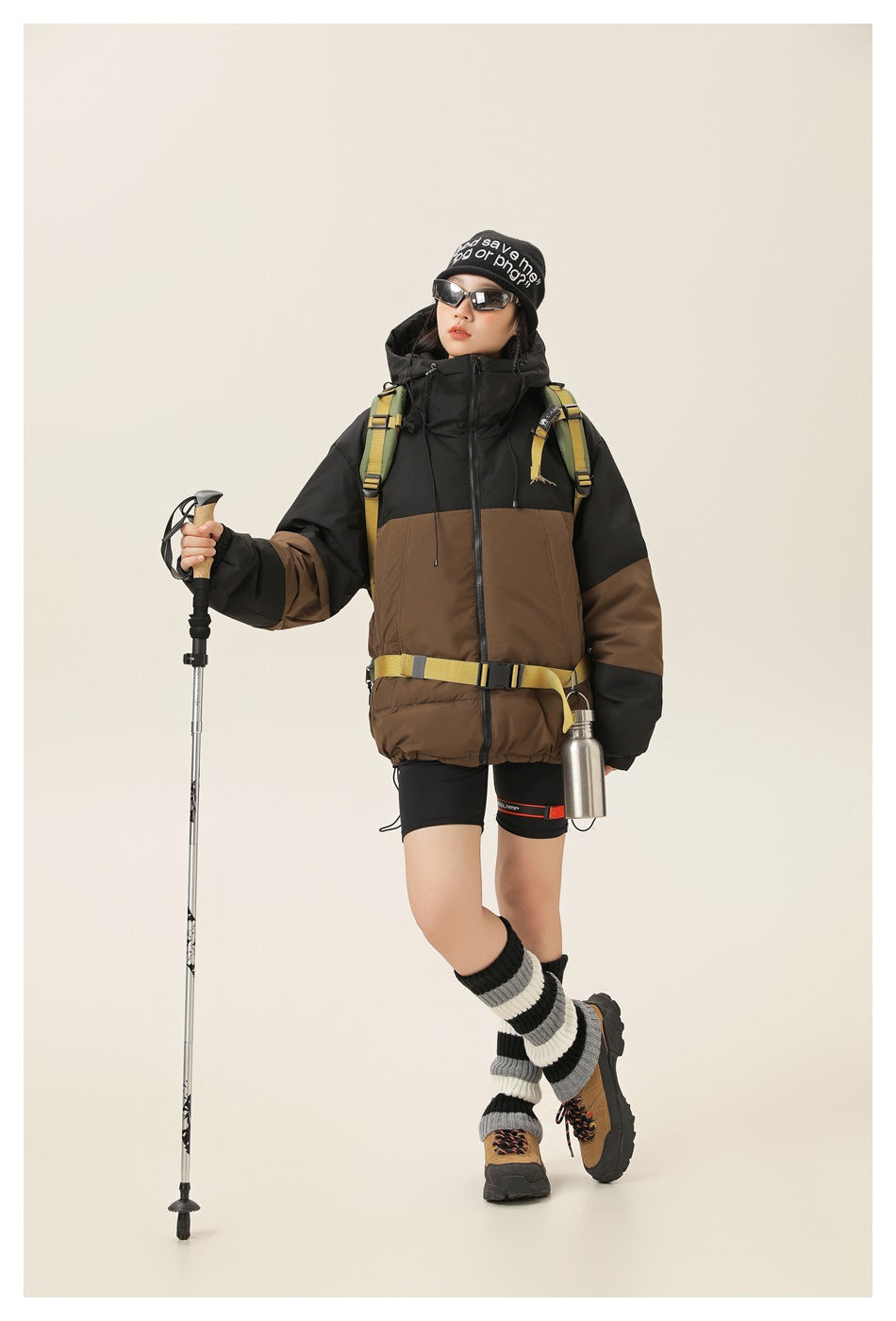 Model in a black and brown hooded puffer jacket with backpack, trekking pole and water bottle