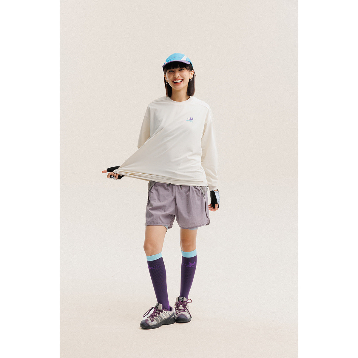 Model wearing an ivory long-sleeve shirt with a small chest emblem and mauve shorts, with a blue cap and tall socks.