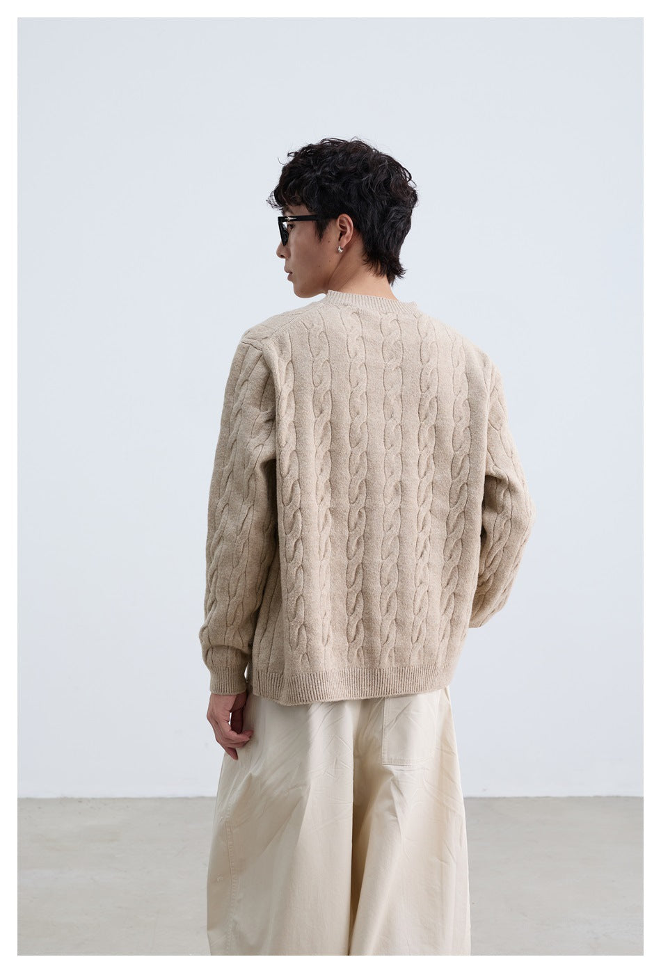Back view of a person wearing a beige cable-knit crewneck sweater with ribbed cuffs and hem.