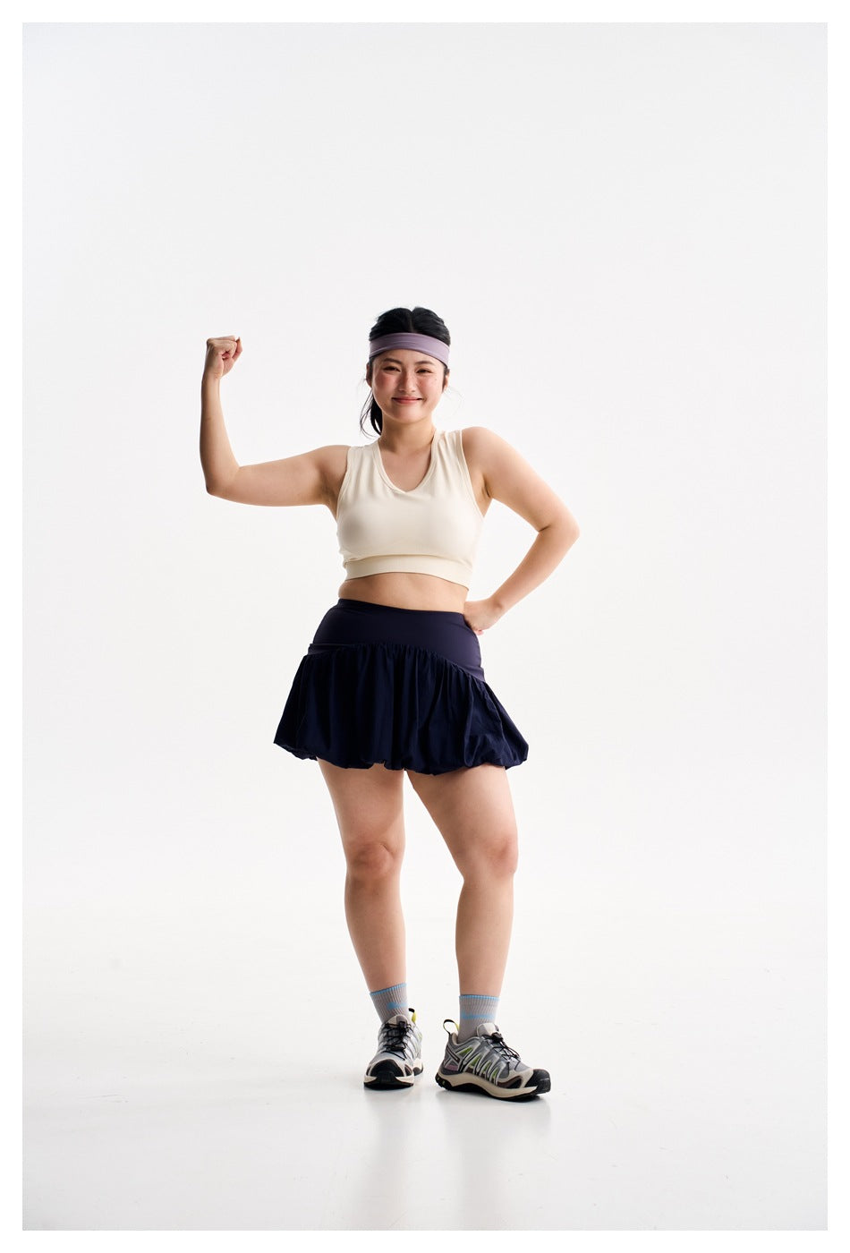 Woman in an ivory crop top and navy bubble-hem athletic skirt, posing with one arm raised.