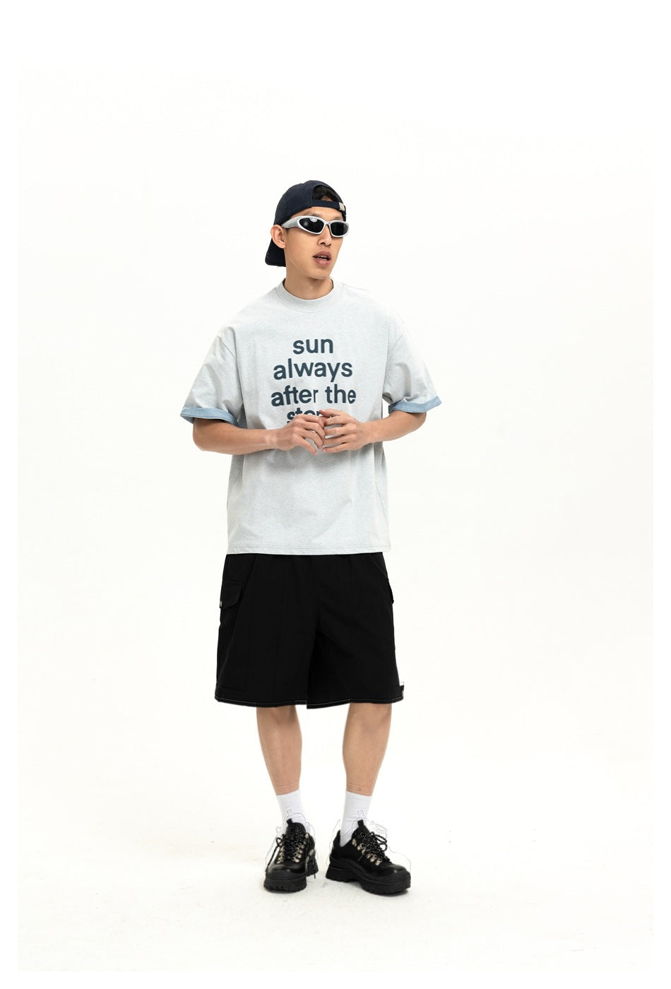 Model wearing a light gray slogan T-shirt with blue sleeve cuffs and black shorts, with a backward cap and sunglasses.