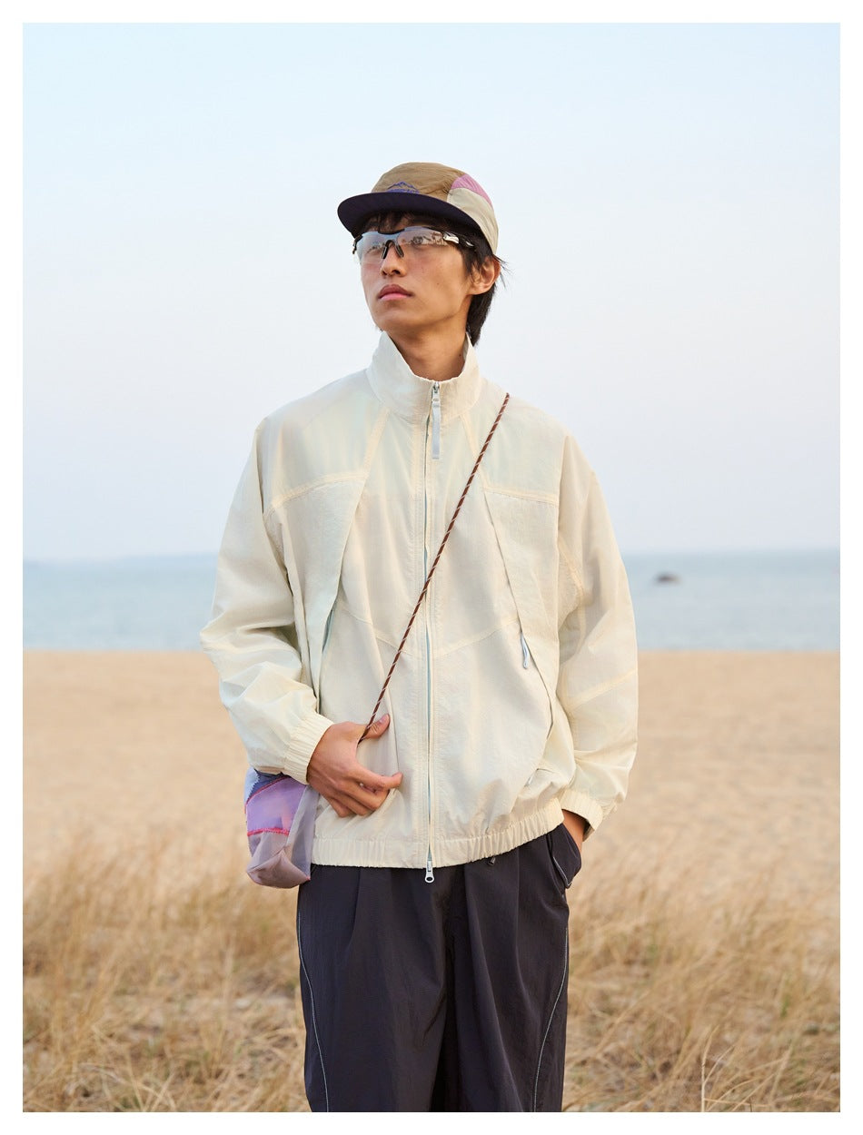 A model wears a cream zip jacket with dark trousers and a color-block cap outdoors.