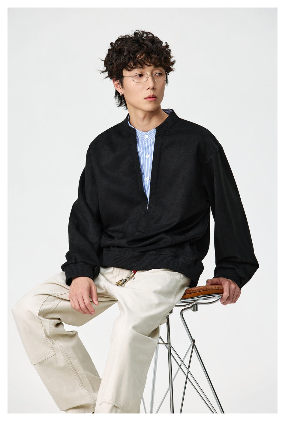 Closer seated view of a model in a black oversized split-neck pullover, blue striped shirt, and cream trousers.