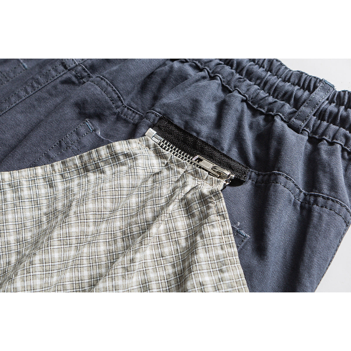 Close-up of slate-blue pants showing the elastic waistband, zipper, and a light plaid fabric panel.
