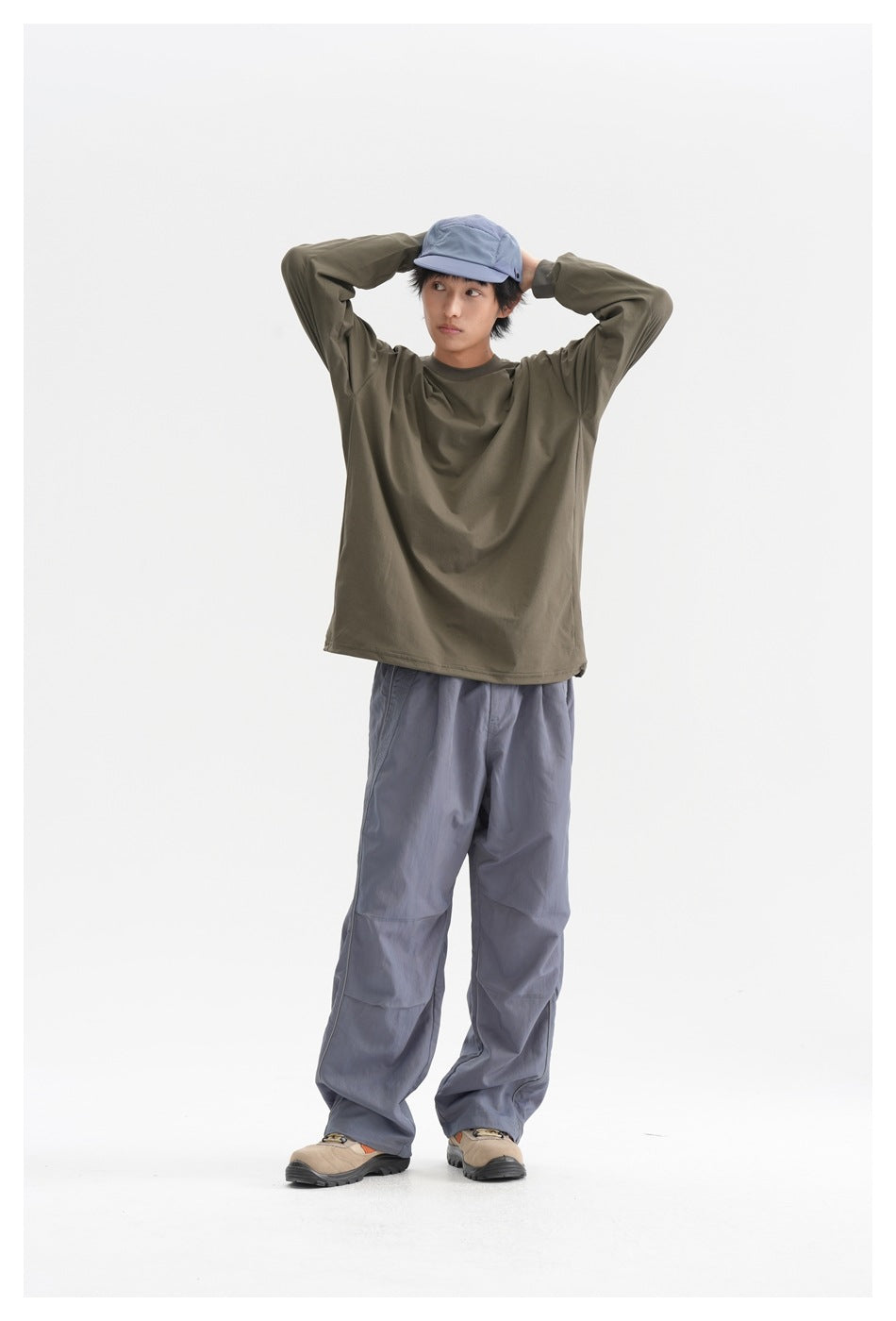 Full-length studio view of a model wearing an olive long-sleeve shirt, slate-blue loose pants, and a blue cap.