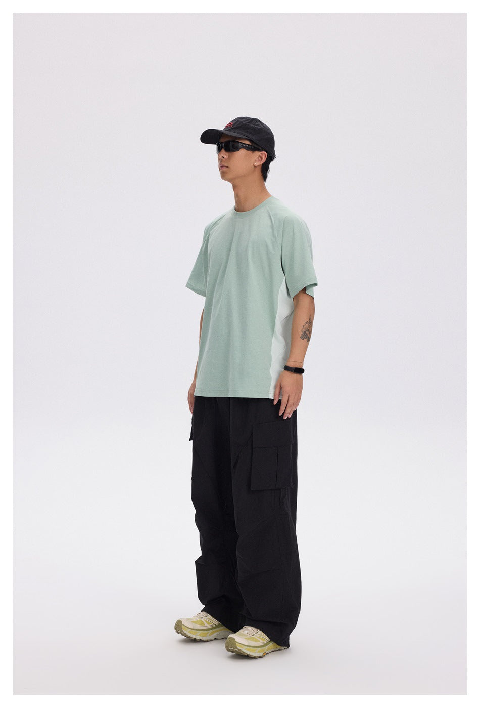 Side view of a model wearing a pale mint oversized T-shirt, loose black cargo pants, a black cap, and sunglasses.