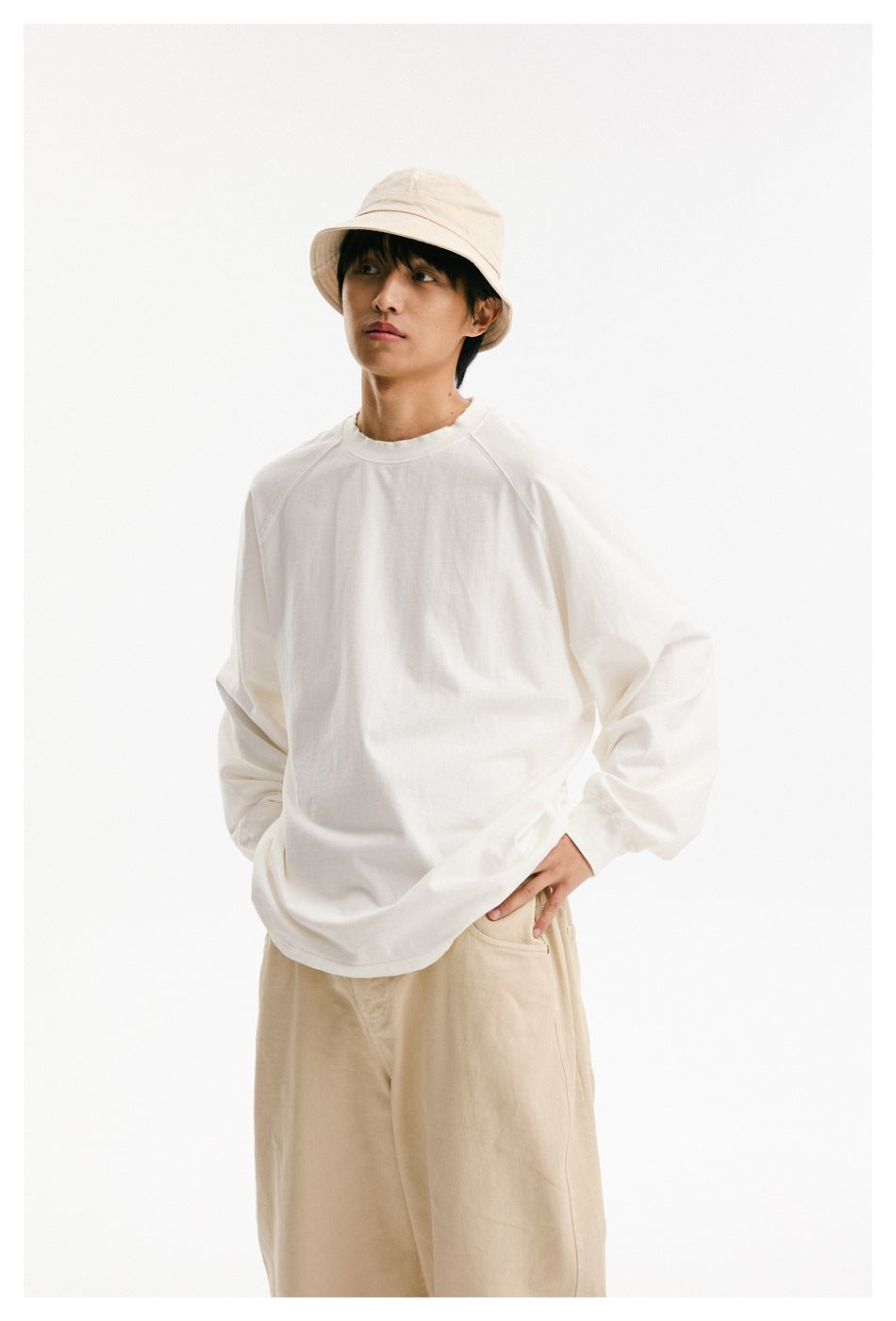Model wearing an oversized white raglan long-sleeve shirt, beige bucket hat, and tan trousers.