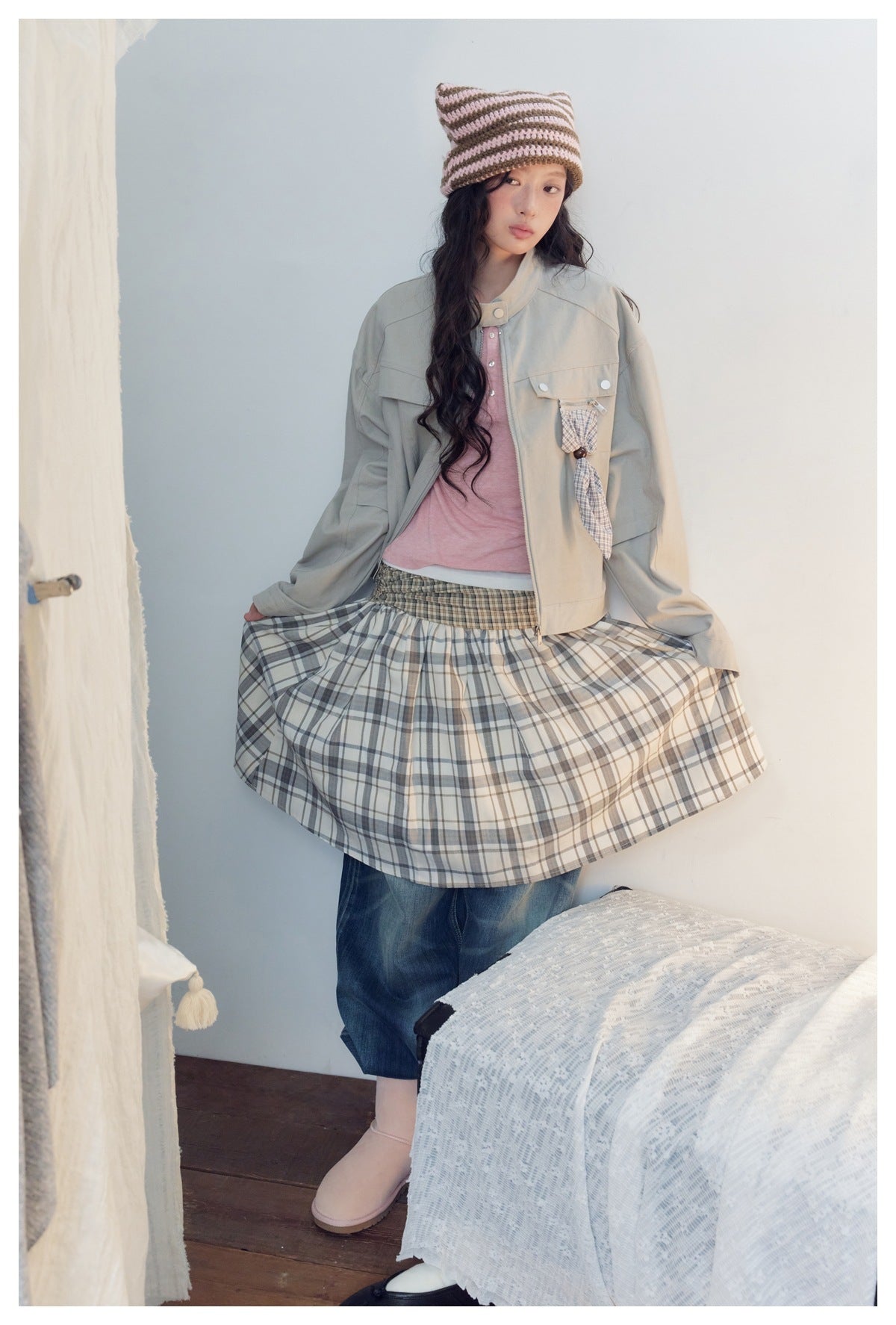 Model wearing a light gray-beige utility jacket with flap pockets and a plaid hanging detail, holding out a plaid skirt.