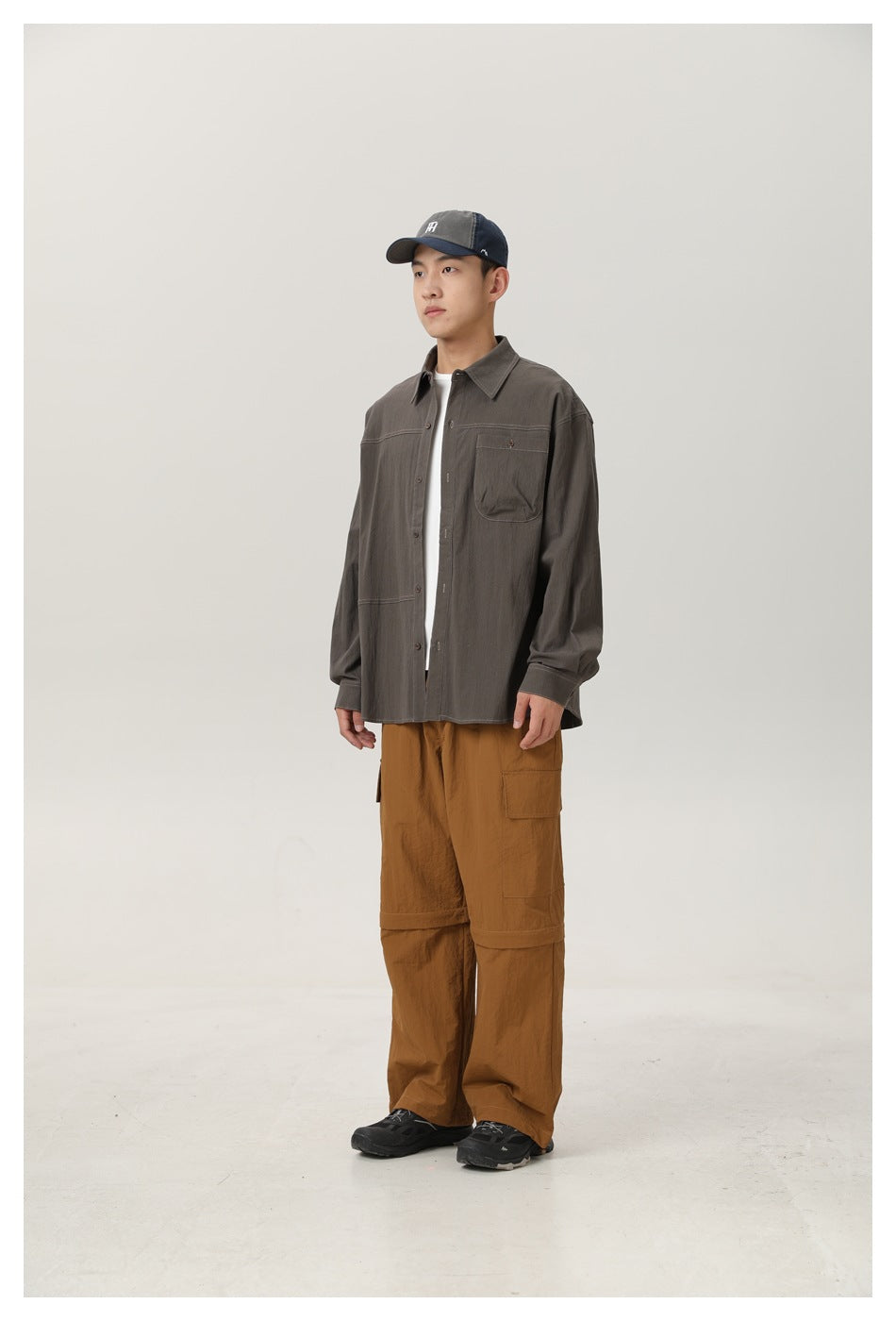 Man wearing open gray-green button-up shirt over a white tee with brown cargo pants and a cap, full-length view
