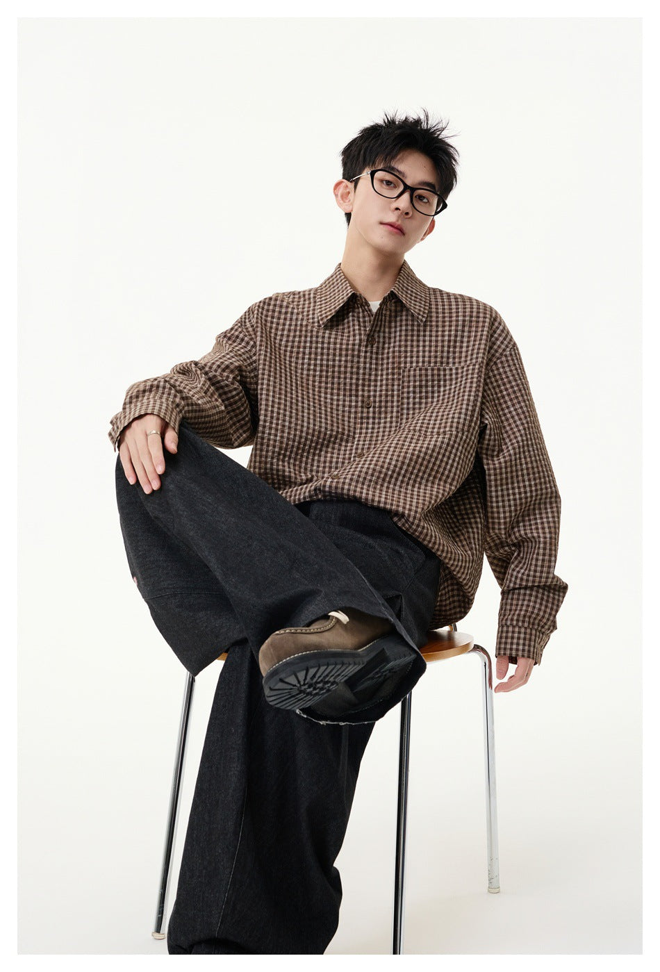 Seated model wearing a brown checked button-up shirt and wide dark trousers.