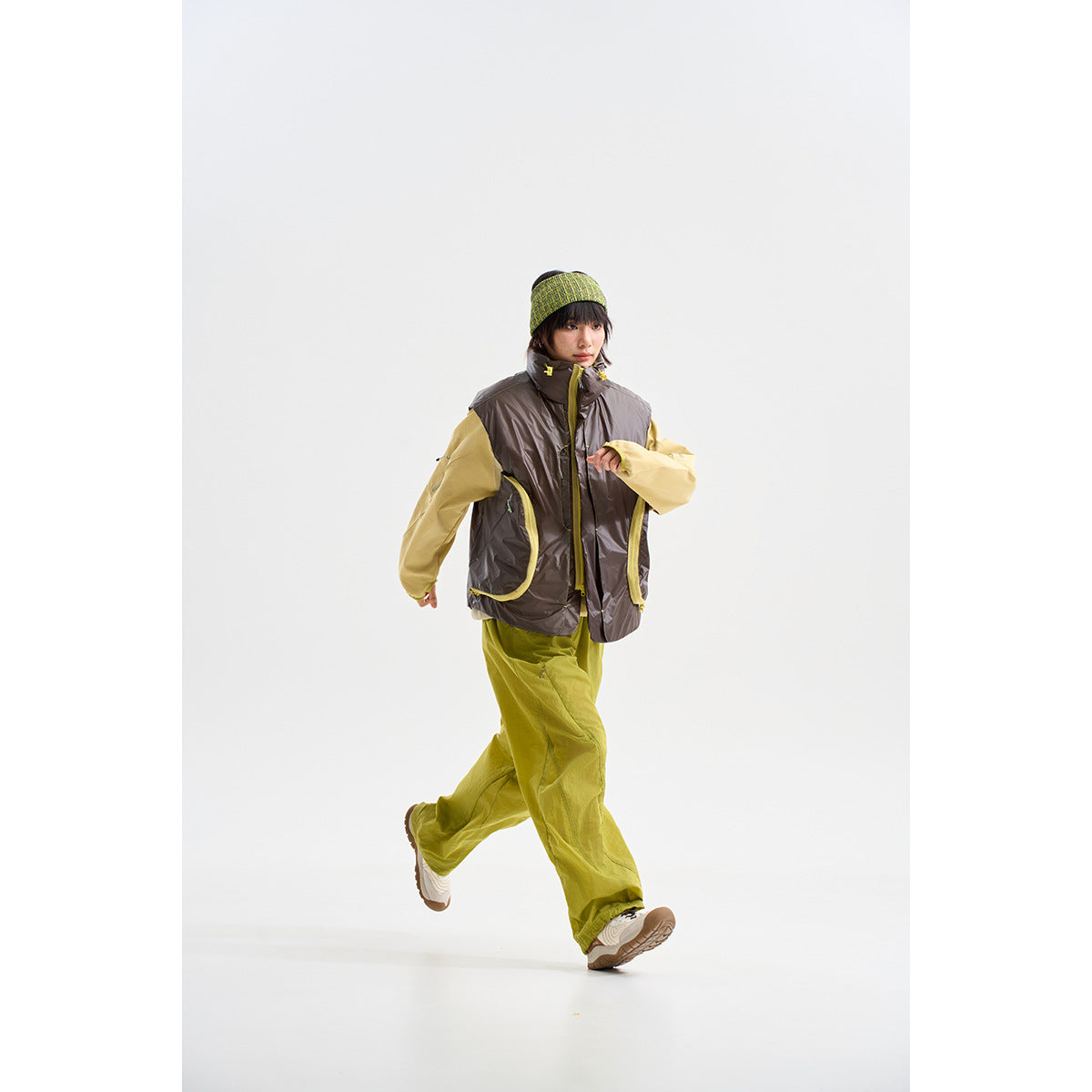 Model in a glossy dark sleeveless vest with yellow trim, pale yellow sleeves, lime trousers and a green beanie.