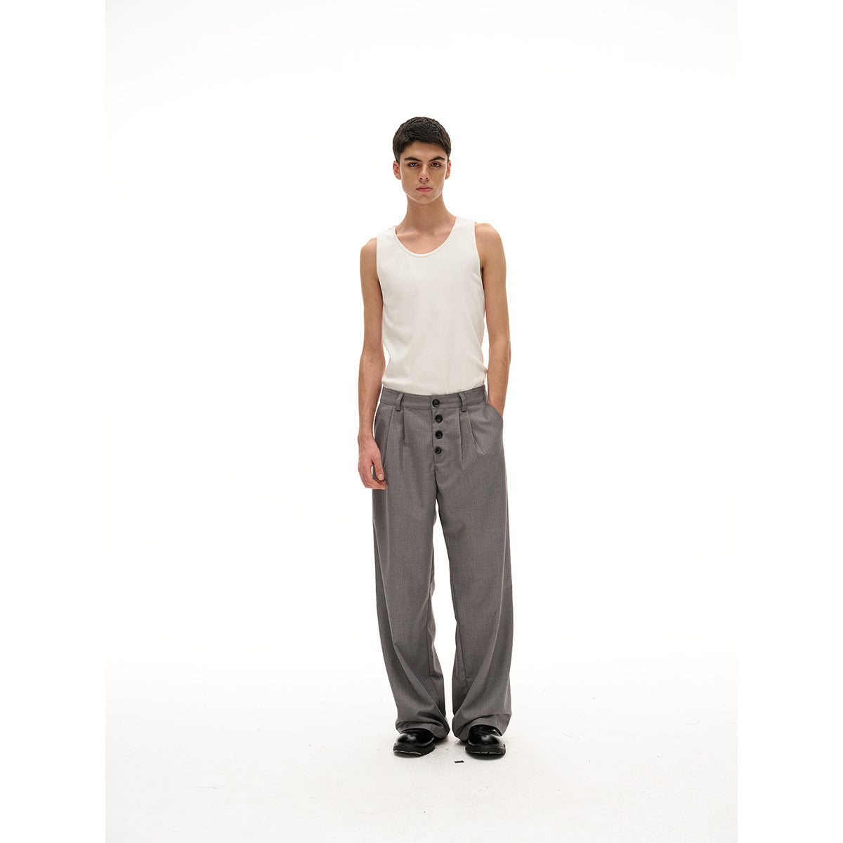 Model wearing a white tank top and loose gray pleated trousers with a four-button front closure, facing forward.