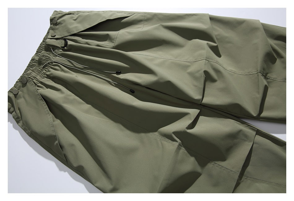 Olive-gray pants laid flat, showing the elastic waist, drawcords, flap pocket and roomy leg panels.
