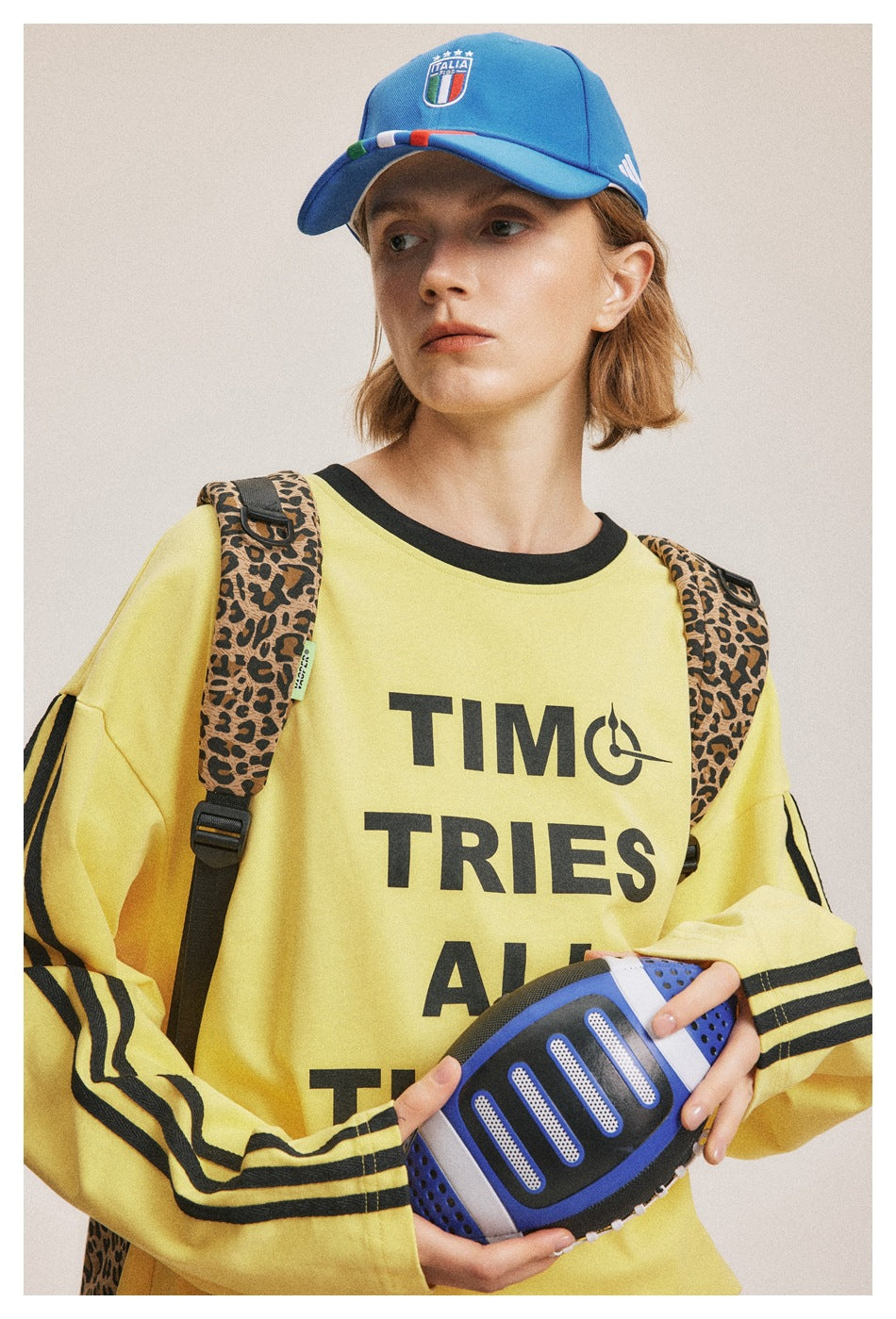 Close-up of a model wearing a yellow slogan sweatshirt with black stripes and a blue cap, holding a ball.
