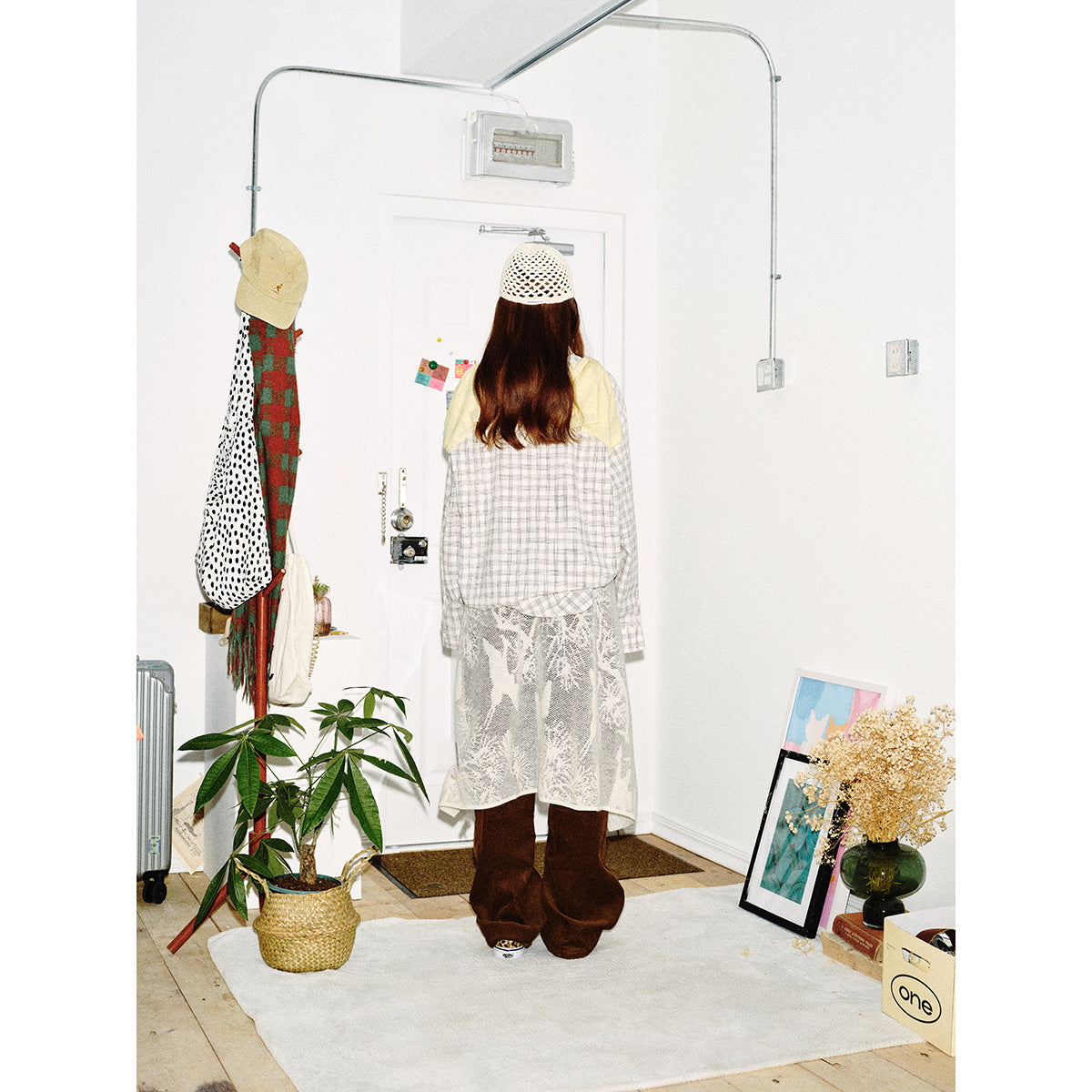 Full-length back view of a model wearing a light plaid shirt, white lace apron, brown pants and crochet cap.