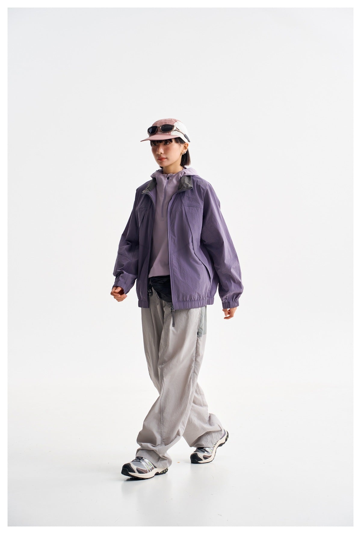 Model wearing a purple zip-front windbreaker over a lavender top with loose light gray pants, shown from a three-quarter