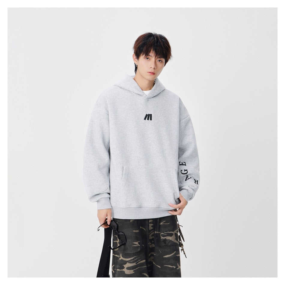 Model wearing a light gray oversized hoodie with a black chest mark and sleeve lettering, shown from the front.