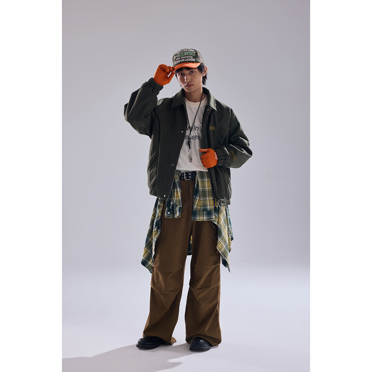 Model wearing a dark olive jacket, white graphic tee, plaid shirt tied at the waist, and brown trousers.