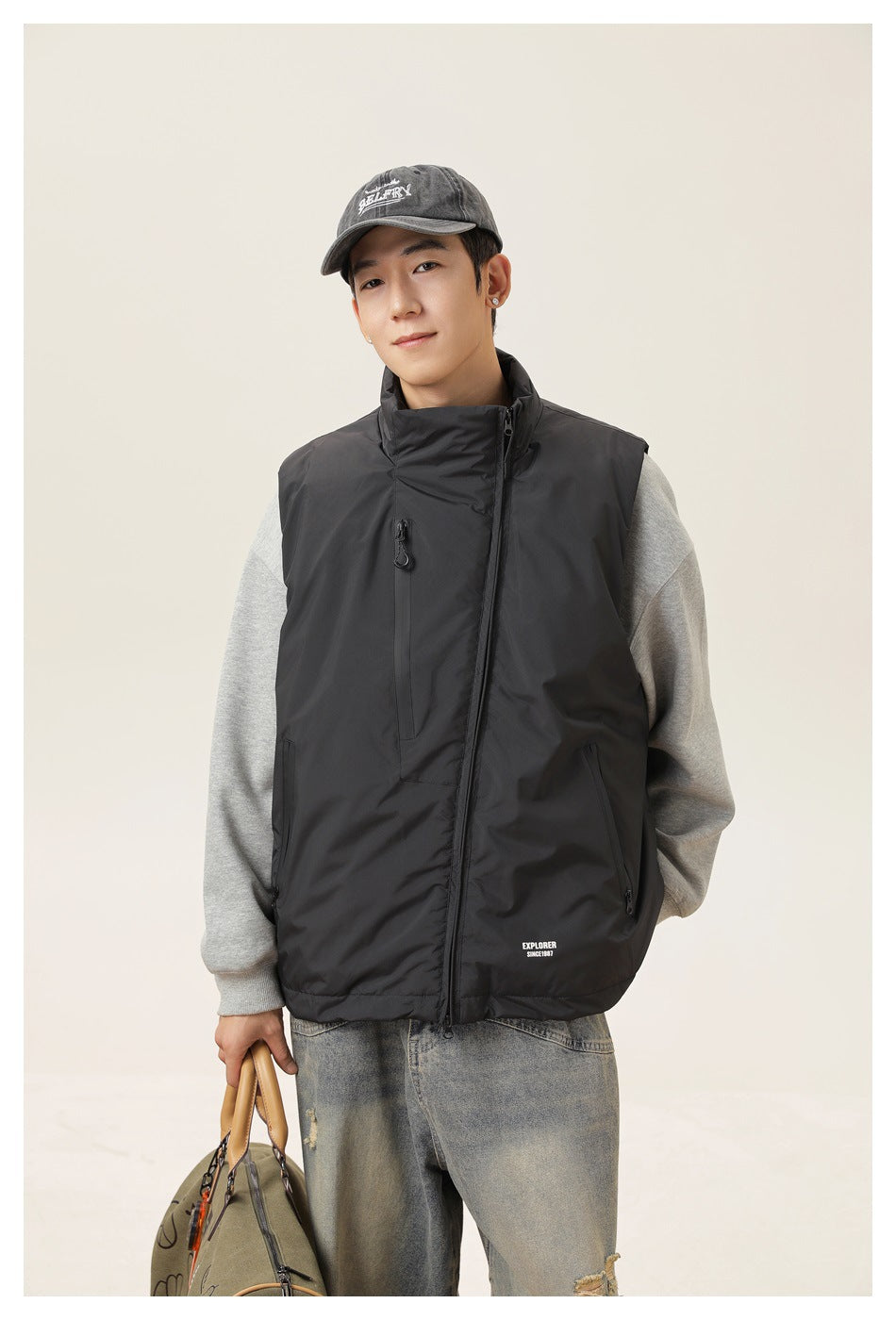 Closer front view of a model wearing a black zip-front vest with a high collar and zip pockets over a gray sweatshirt.