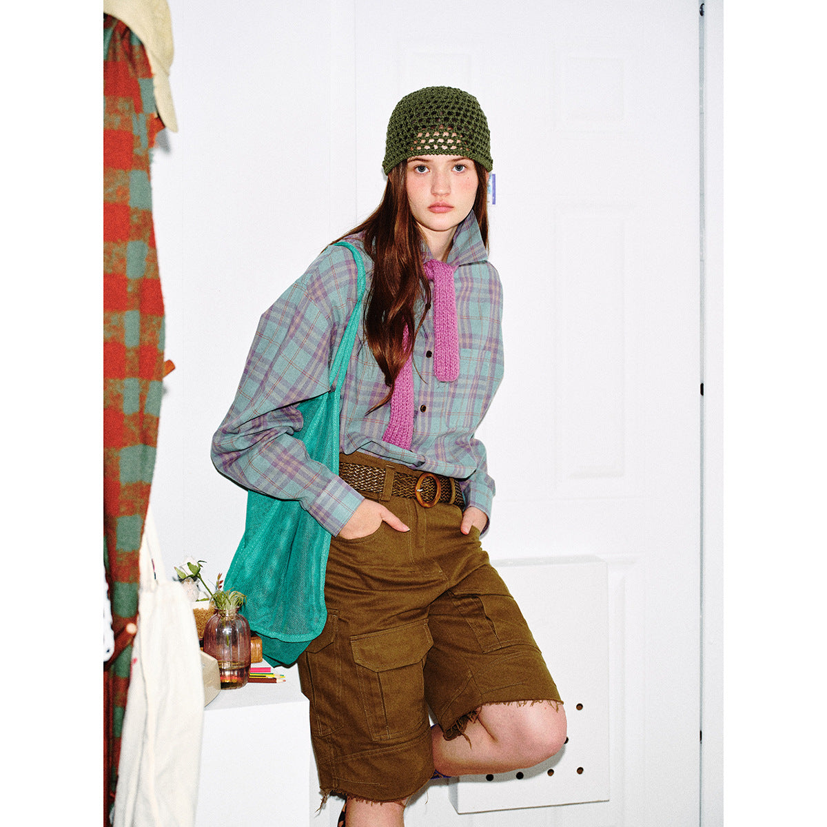 Model wearing a pastel plaid button-up shirt with a pink tie detail, brown cargo shorts, and a green crochet cap.