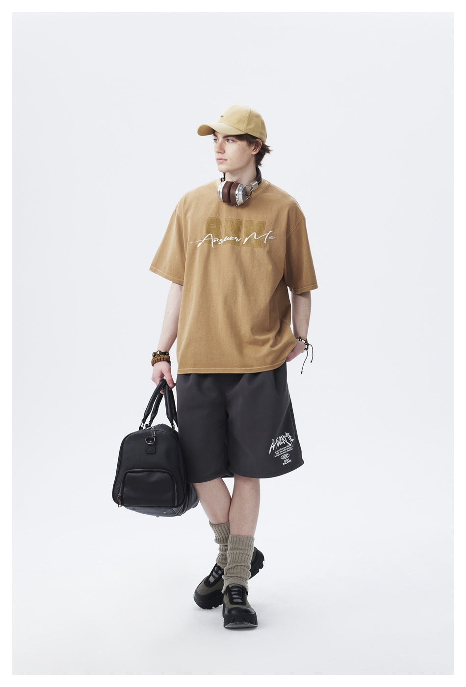 Full-length model wearing a tan graphic T-shirt, charcoal shorts, and a matching beige cap.