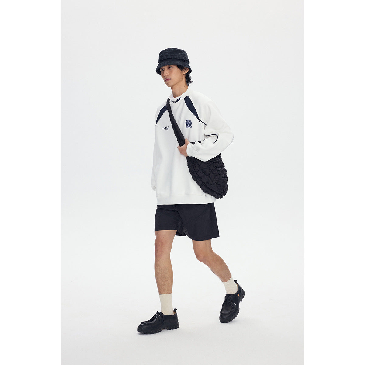 Model wearing a white sweatshirt with navy accents, black shorts, and a black shoulder bag.
