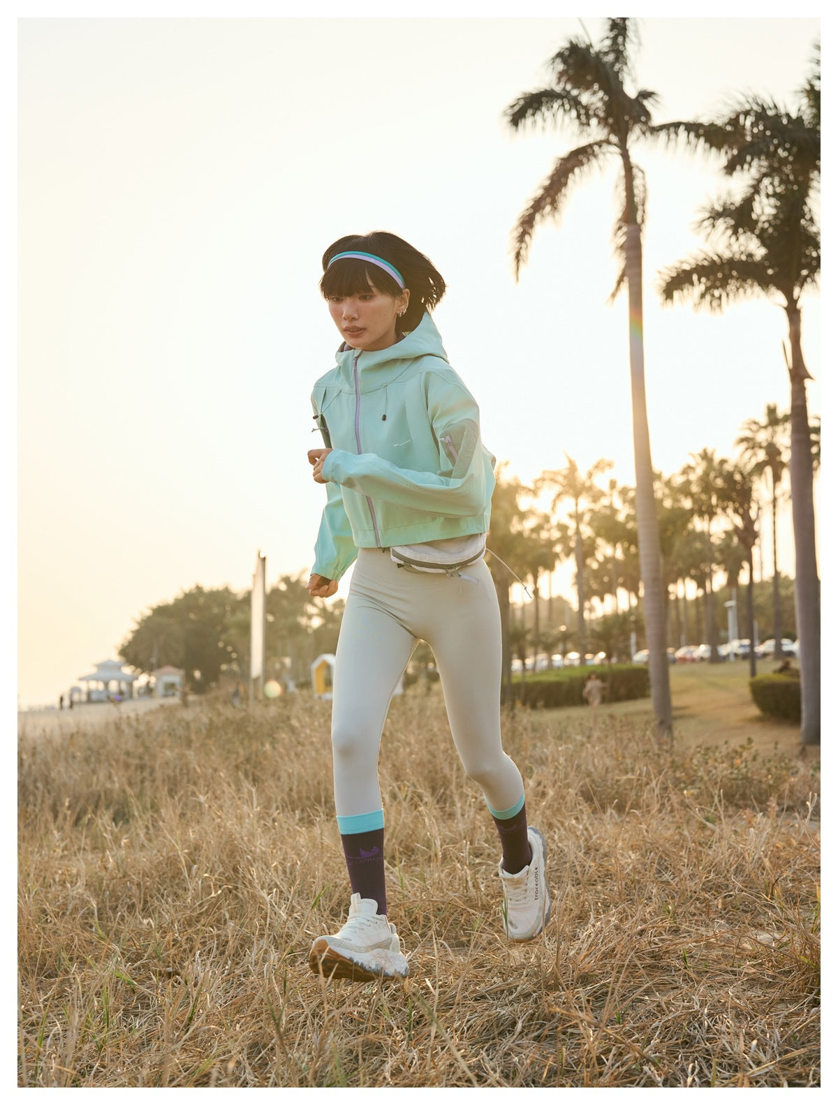 Model walking in a mint cropped hooded jacket, pale leggings and purple socks beside a beach.