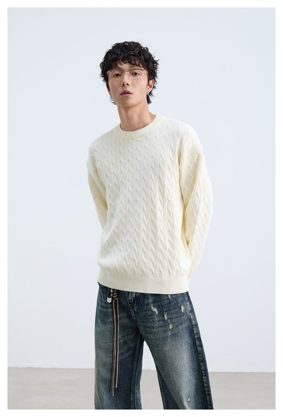 Model wearing an ivory cable-knit crewneck sweater with dark distressed jeans, shown standing from the front.