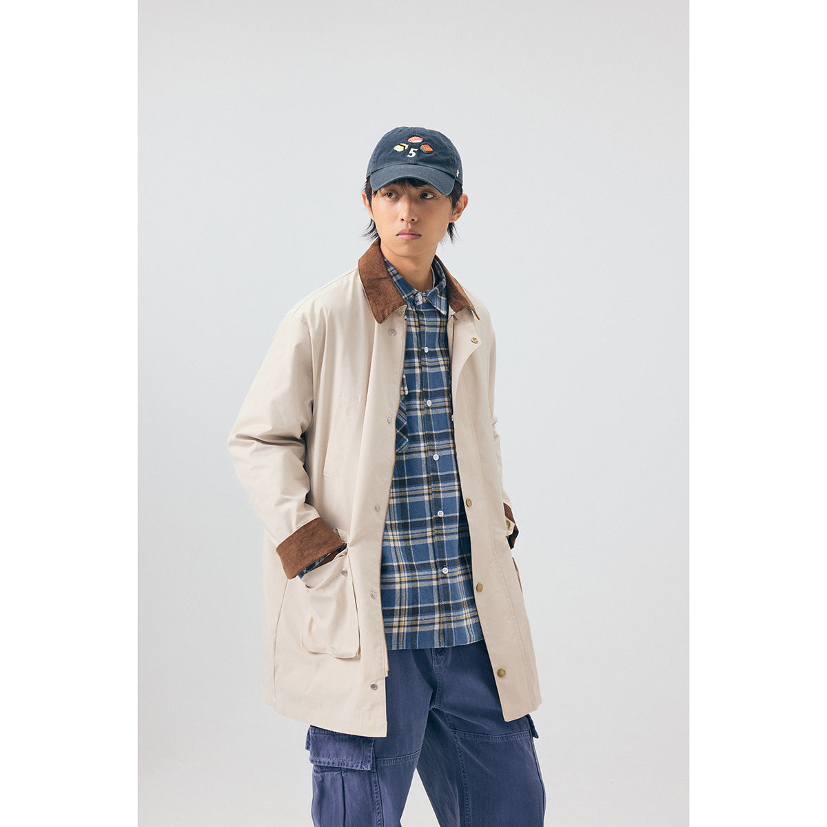 Model wearing a light beige coat with a brown collar and cuffs, layered over a blue plaid shirt.