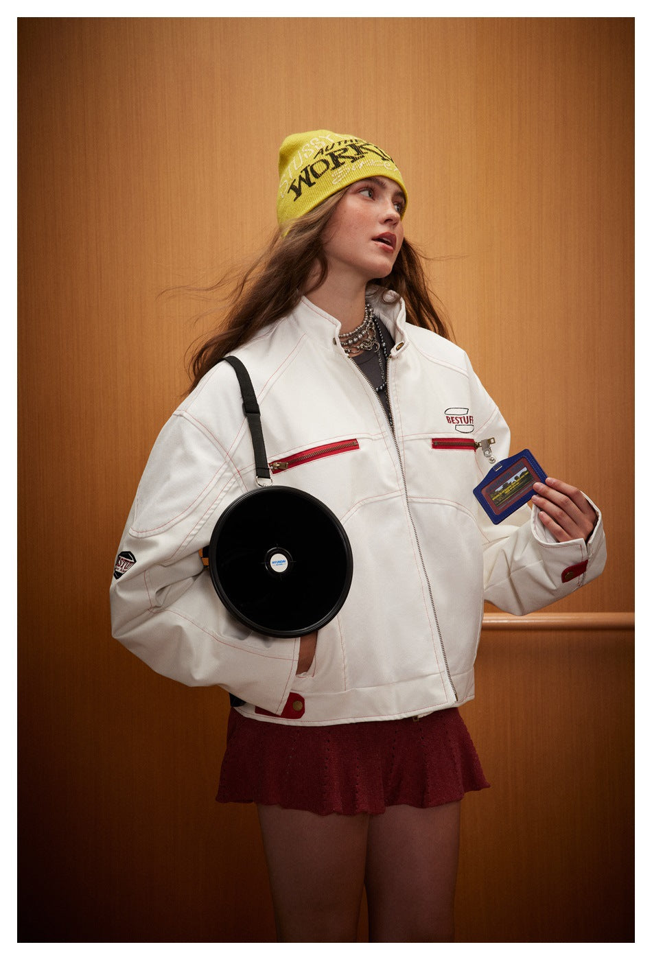 Model wearing an oversized white jacket with red zip pockets, paired with a burgundy skirt and yellow beanie.