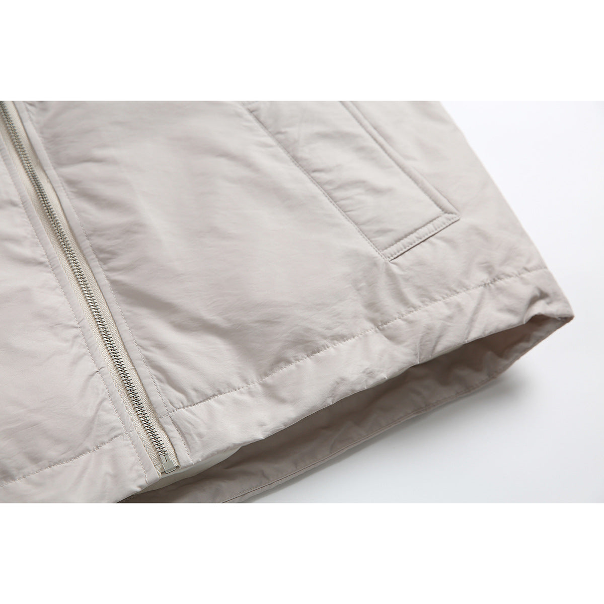 Close-up of a light beige jacket's front zipper, pocket seam, and bottom hem.