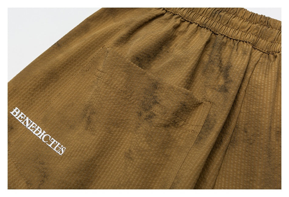Close-up of brown shorts with a stitched pocket and white embroidered BENEDICTUS lettering.