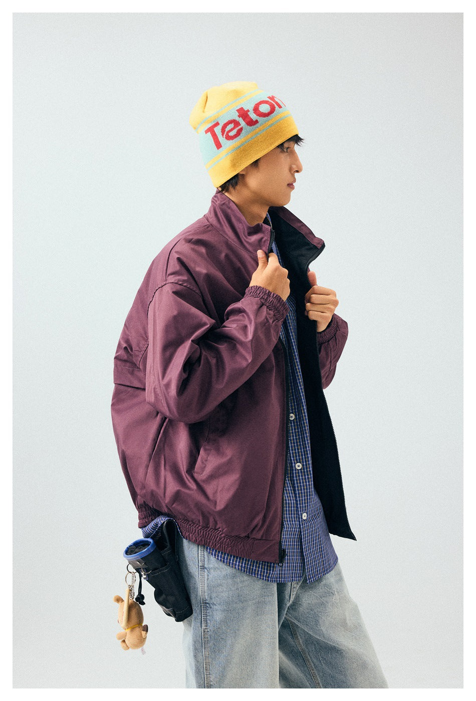 Side view of a model wearing an open plum jacket over a blue checked shirt, with a yellow graphic beanie.