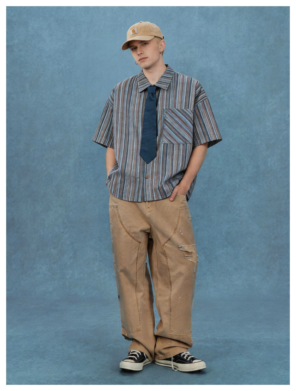 Man in navy striped short-sleeve shirt with tie, hands in pockets, tan cap and wide-leg pants