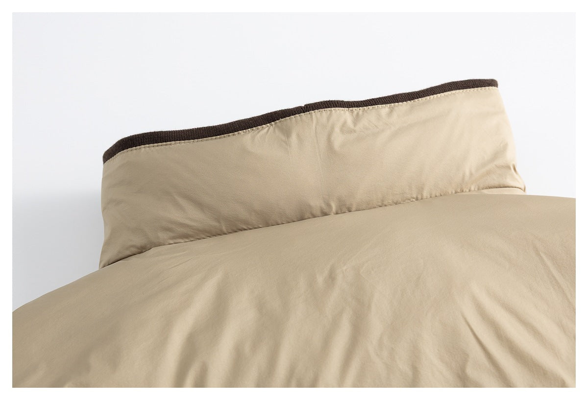 Close-up of beige bedding with a pillow edged in dark brown piping behind the comforter.