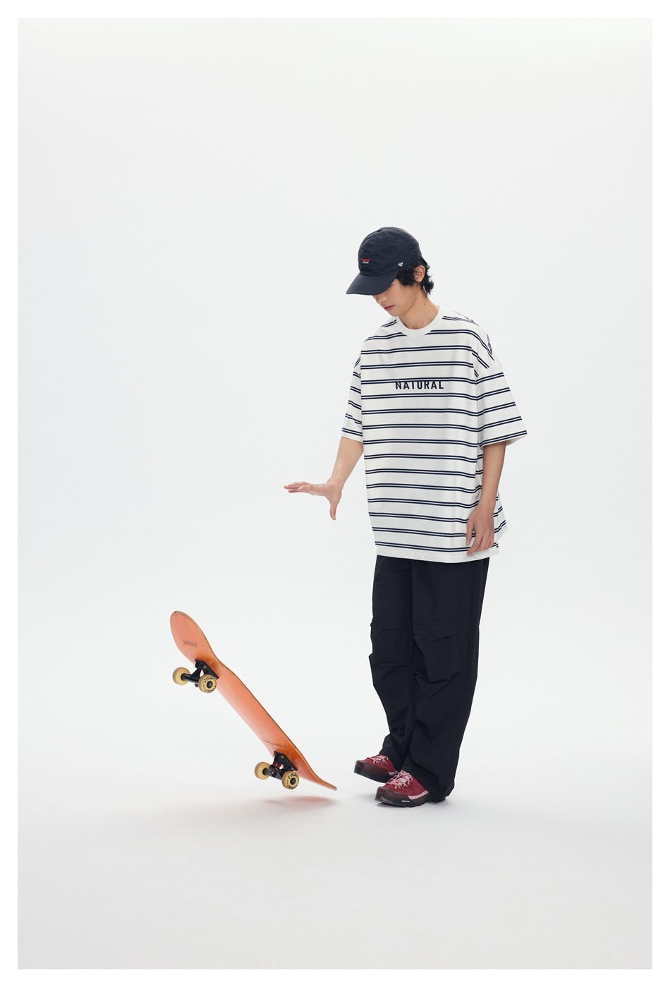 Model wearing a white T-shirt with thin navy stripes and NATURAL text with loose black pants, beside a tilted skateboard.