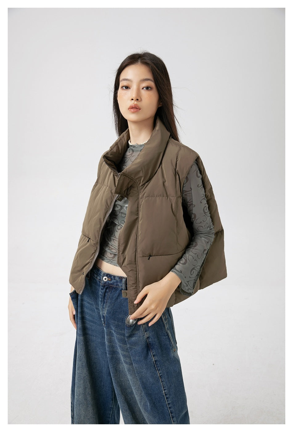 Front view of a model wearing a taupe quilted sleeveless vest with a high collar over a patterned gray top.