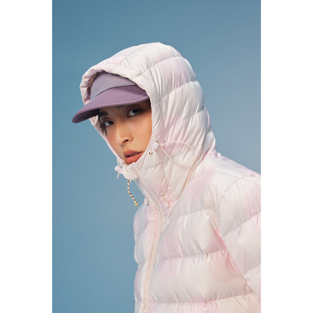 Close-up of a model wearing a pale pink hooded puffer jacket and lavender cap against a blue background.