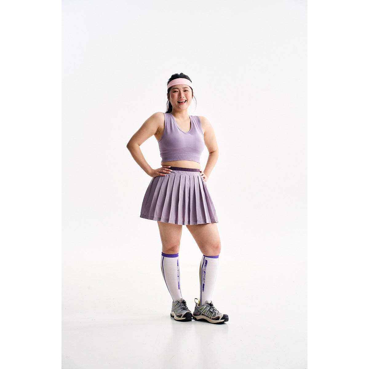 Model wearing a lavender cropped sports bra and pleated skirt with a pink headband and knee-high socks.
