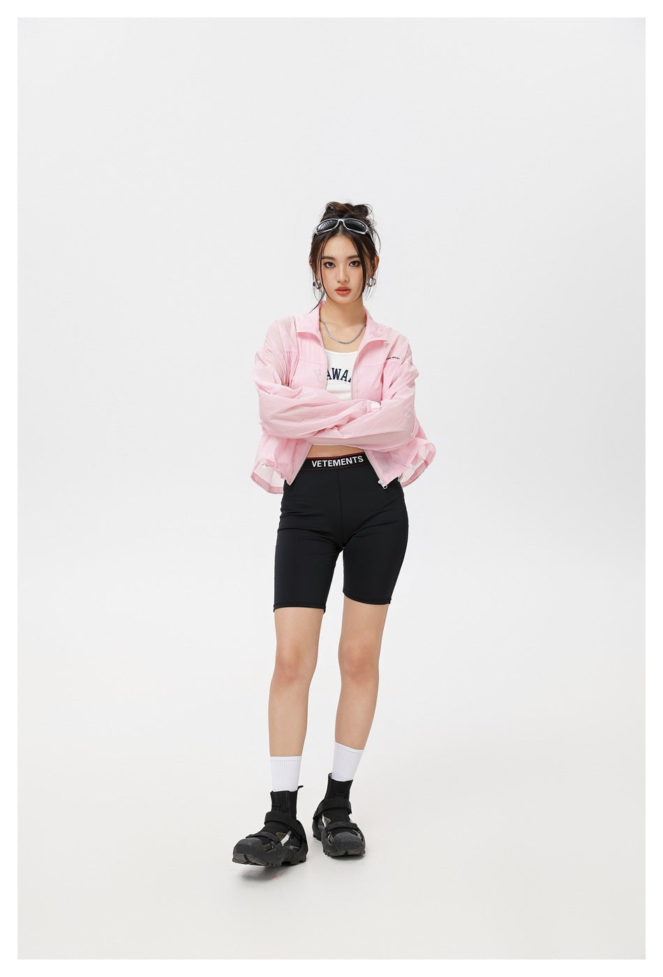 Model wearing a light pink zip-front jacket with a stand collar and black cycling shorts; her arms are crossed.
