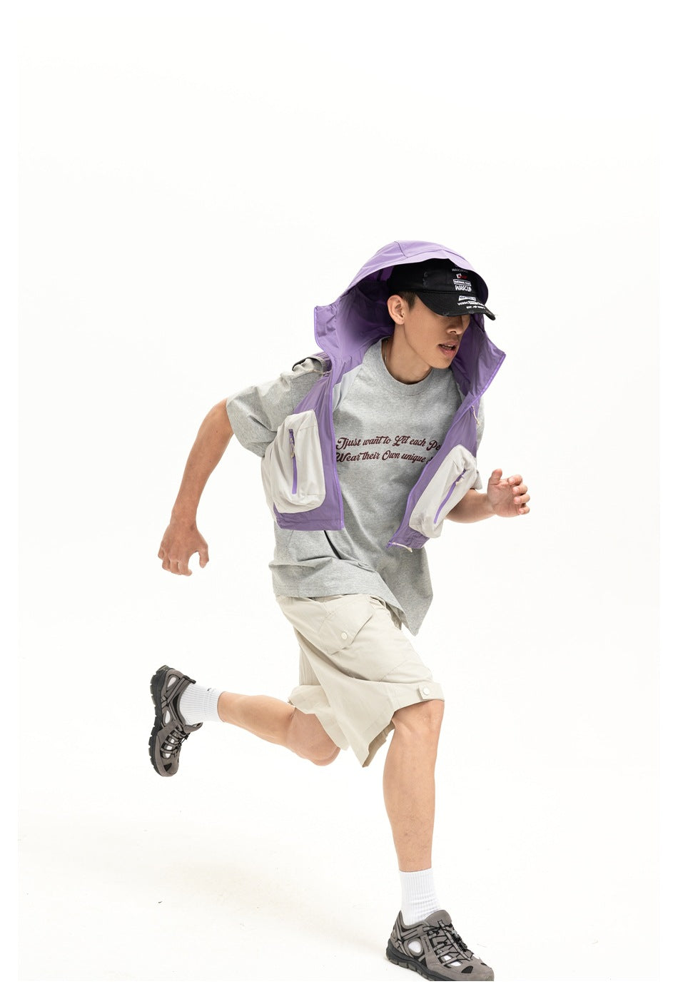 Model in a purple hooded utility vest with white pockets, gray graphic T-shirt, and cream cargo shorts, shown in motion.
