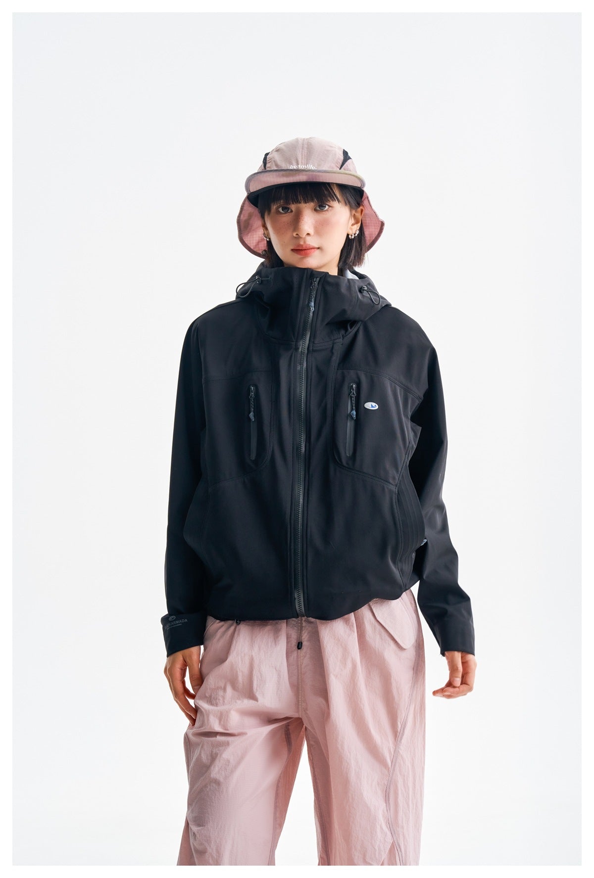 Front view of a model in a zipped black hooded jacket with two vertical chest pockets and pink trousers.