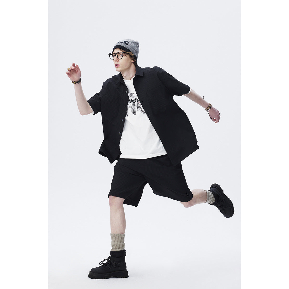 Model in a black short-sleeve button-up shirt and matching shorts over a white graphic tee, shown mid-stride.