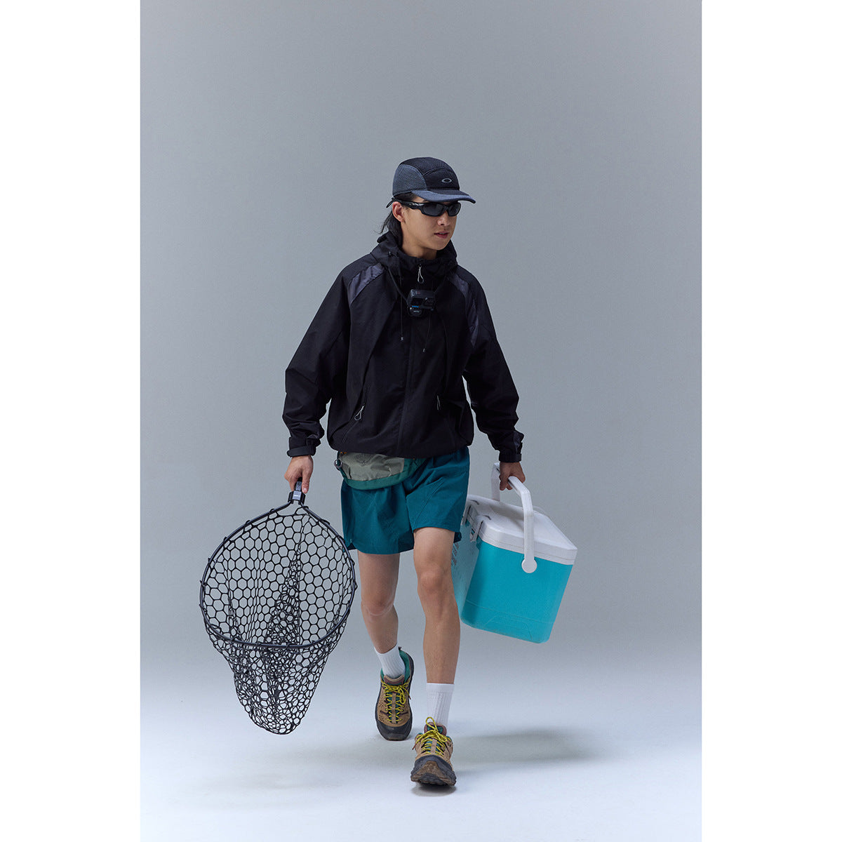 Man wearing a black hooded jacket and teal shorts, shown carrying fishing gear.