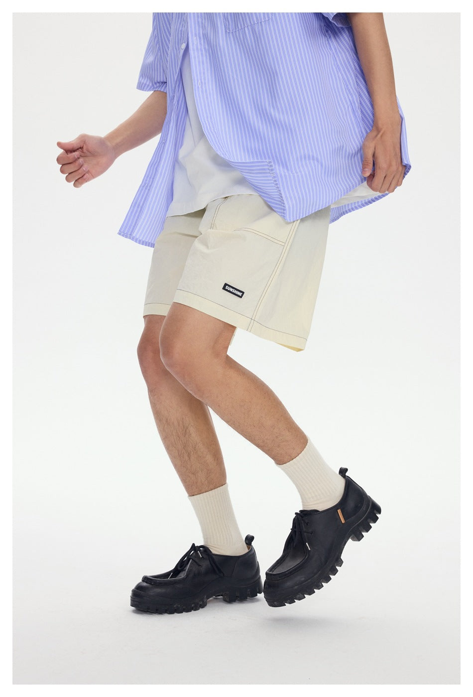 Model wearing cream knee-length shorts with contrast stitching and a small black label, paired with a blue striped shirt.