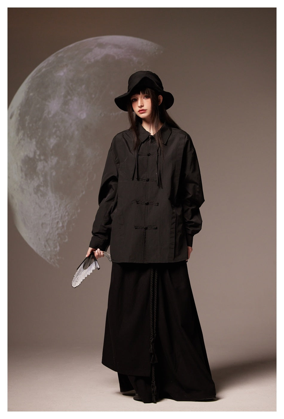 Model wearing a black frog-closure jacket, wide-brim black hat, and long black skirt against a moon backdrop.